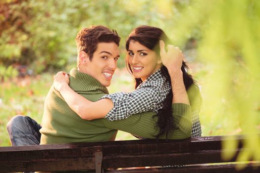 Photo of a young heterosexual couple hugging sitting on a park bench, behind their back looking at the camera with a smile on their faces.