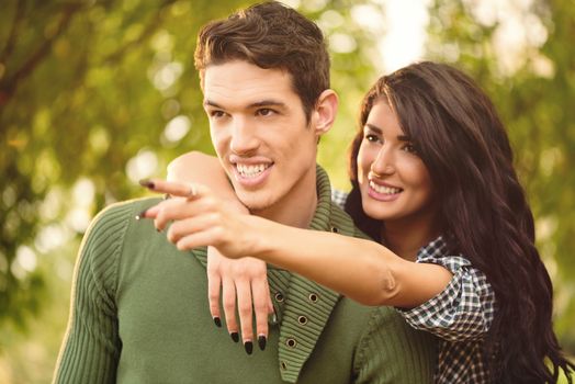 Young heterosexual couple in love, girl standing behind the guy with her hand slung over his shoulder while with the other hand pointing to something with outstretched finger. In the background is greenery.