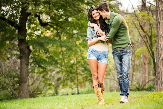 Young cheerful couple, attractive brunette and her handsome boyfriend, embracing and smiling walking through the park.