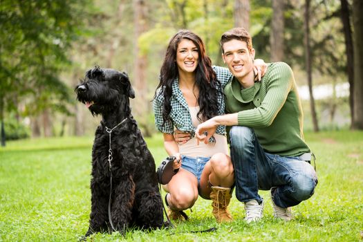 Young attractive girl crouches in the park with her boyfriend next to the dog, a black giant schnauzer. With a smile looking at the camera.