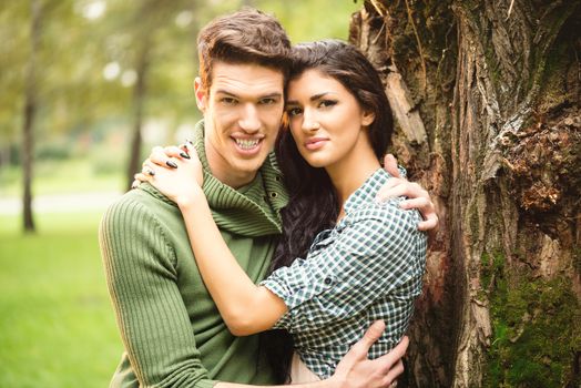 Young beautiful guy and girl are embraced beside the tree and with a smile looking at camera.