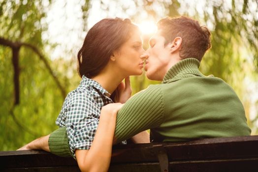 Young heterosexual couple photographed from behind while sitting on a park bench, kiss, illuminated by the rays of the spring sun.