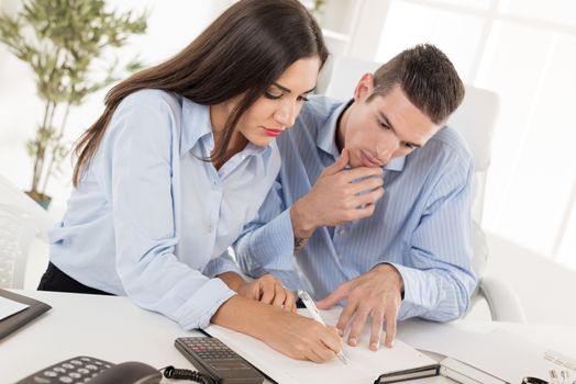 Two young business people, man and woman sitting in office at an office desk and looking at the organiser in which businesswoman writes.