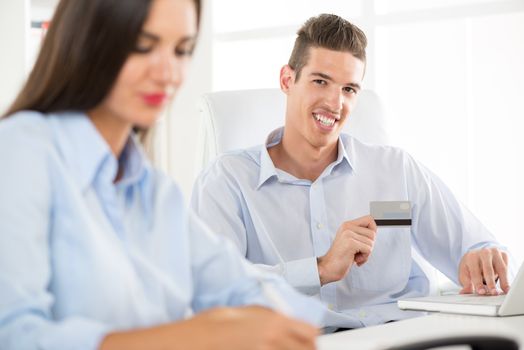 Young business people, man and woman, sitting in an office, a young businessman who is in the foreground holding a credit card and with a smile looking at the camera.