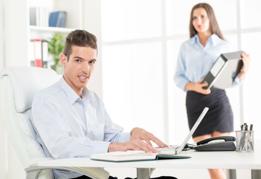 Young businessman sitting in office chair in front of a laptop, with a smile looking at the camera in the background is a young woman holding a binder.