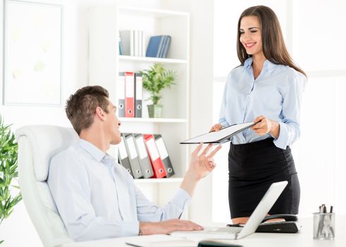 Young business people in the office, a woman standing next to office desk with a smile on her face gives the folder to young businessman who sits in office chair.