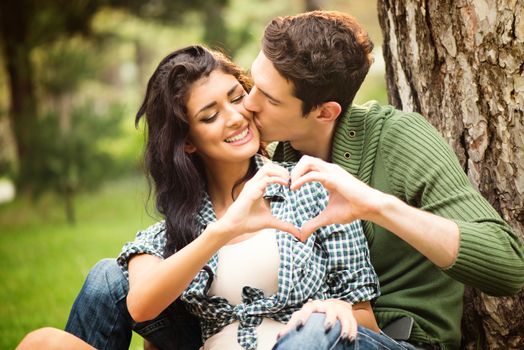 Young heterosexual couple sitting against a tree in the park, boy kissing girl, while touching with fingers forming a heart shape.