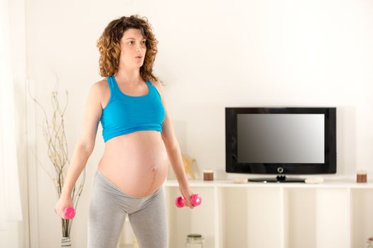 Pregnant woman in sports clothes, standing in the living room, holds the dumbbells and doing breathing exercises.