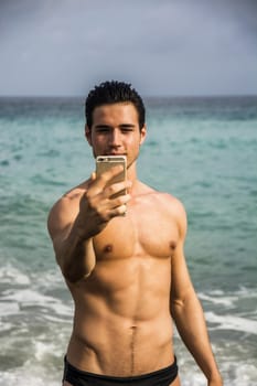 Side View of a Shirtless Young Man Taking Selfie Photos at the Beach While Standing Under the Sun.