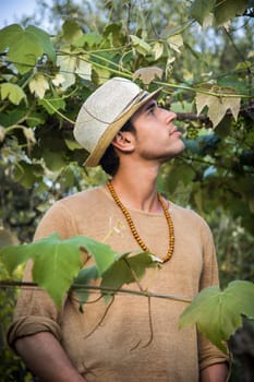 Side view of handsome young man in hat toching vine leaves in garden in sunlight