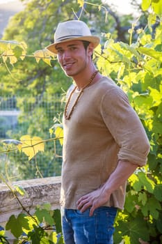 Side view of handsome young man in hat toching vine leaves in garden in sunlight