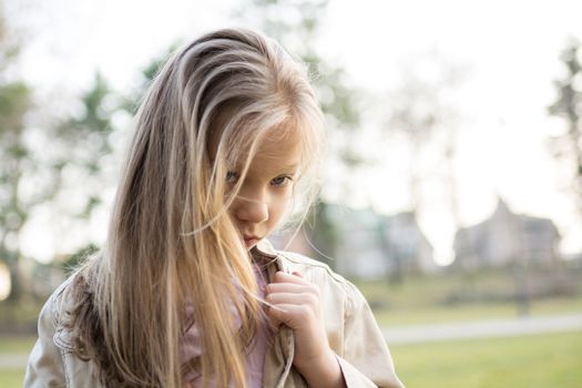 Close-up of a little girl with long hair standing alone outside with a sad expression on her face looking at the camera.