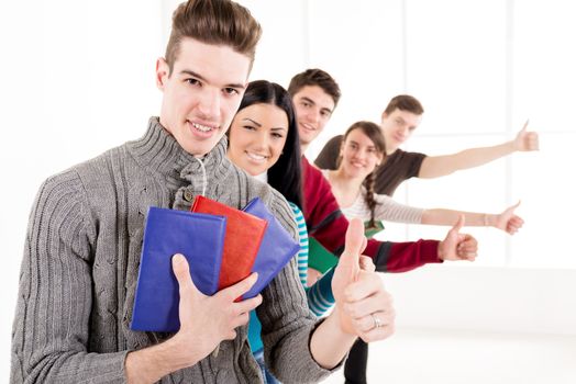 Group of cheerful students with books standing In A Row with thumb up in school hall and looking at camera.