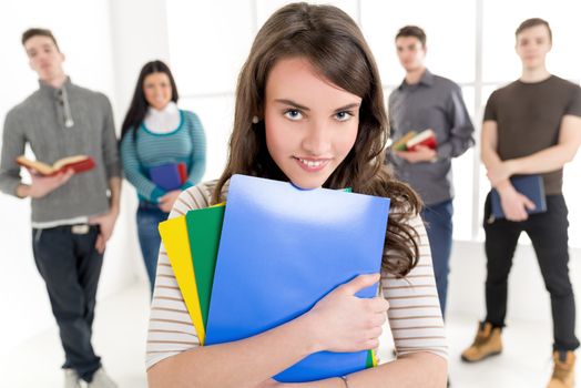 A beautiful smiling girl with books is standing in the foreground. A group of her friends is behind her. Looking at camera.