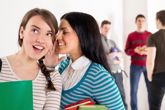 A beautiful teenage girl whispering with her cute friend. They is standing in the foreground. A group of men student is behind them and talking.