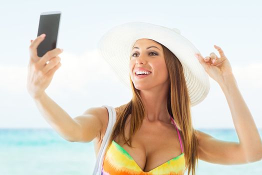 Attractive girl in bikini with Sun Hat taking a selfie on the beach.