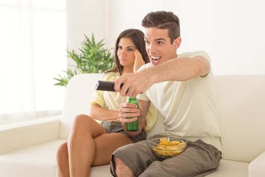 Young man sitting on sofa and watching football on tv. His girlfriend is behind, frowning because and bored.