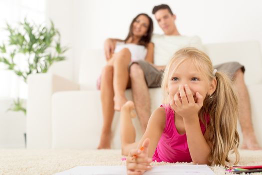 Happy family at home. Mother and father sitting, their cute little girls lying down and drawing with colored pencils. Focus On little girl.