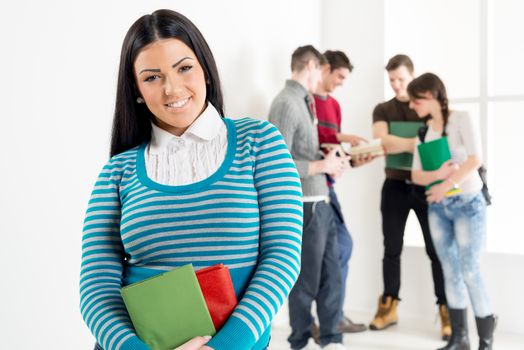 A beautiful smiling girl with books is standing in the foreground. A group of her friends is behind her. Looking at camera.