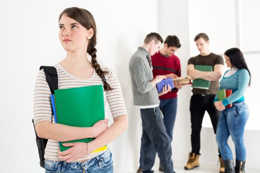 A beautiful smiling girl with books is standing in the foreground. A group of her friends is behind her. 