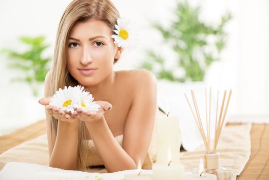 Cute young woman enjoying during a skincare treatment at a spa. She is smiling the camera and holding white flowers in her hands. 