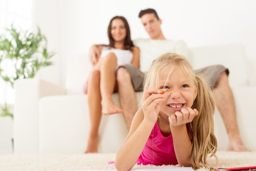 Happy family at home. Mother and father sitting, their cute little girls lying down and drawing with colored pencils. Focus On little girl.