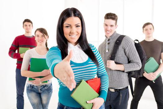 A beautiful smiling girl with books is standing in the foreground and offering her hand as if to shake hands. A group of her friends is behind her. Looking at camera.