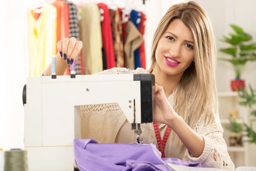 Young beautiful woman sitting in front of a sewing machine on which sets the thread, in the background you can see hanging garments. Looking at camera.