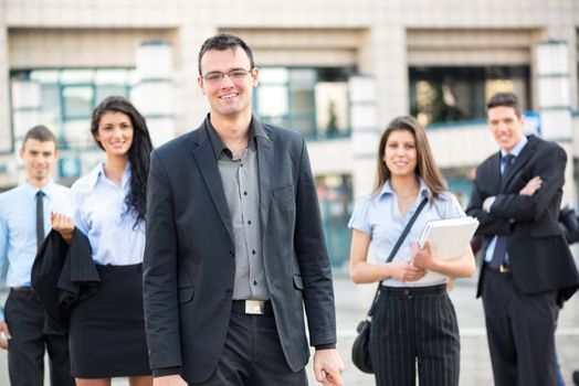 Young businessman, elegantly dressed, standing proudly with his team front of office building.