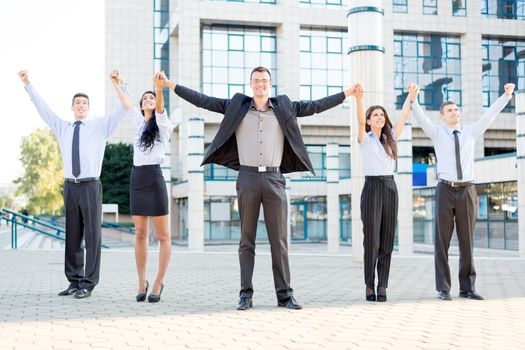 Small group of business people outside their company, holding hands raised celebrate business success.