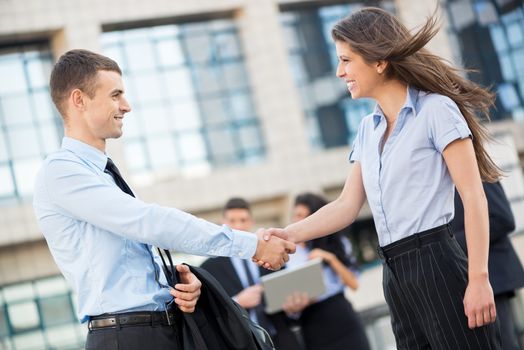 Young businesswoman and businessman shaking hands in front of the company while in the background standing theirs of the business team.