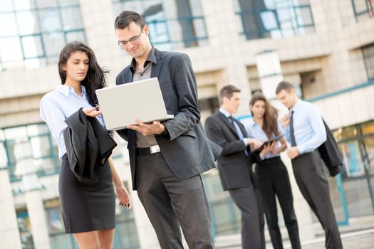Young businesswoman talking to his business partner in front of office building holding a laptop while in the background see the rest of the business team.