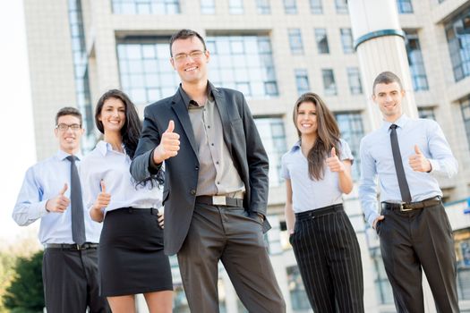 Young business team, elegantly dressed standing outside in front of office building with thumbs up. Looking at camera.