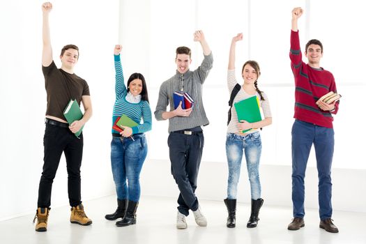 Group of cheerful students with book's standing with arms raised in a fist in school hall and looking at camera.