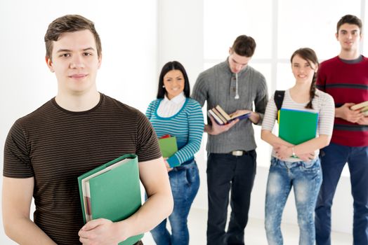 A beautiful smiling young man with book is standing in the foreground. A happy group of his friends is behind him. Looking at camera.