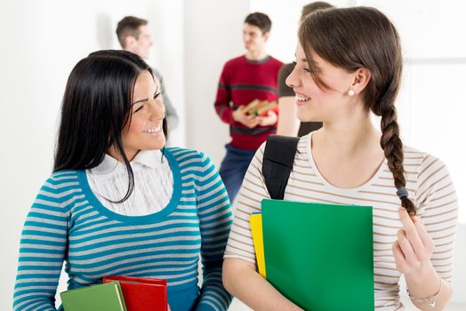 A beautiful two smiling girl with book's is standing in the foreground and talking. A group of men student is behind them.