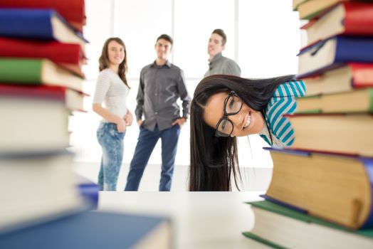 Portrait of a beautiful smiling girl with glasses stick out behind the many book's in the foreground. Looking at camera.