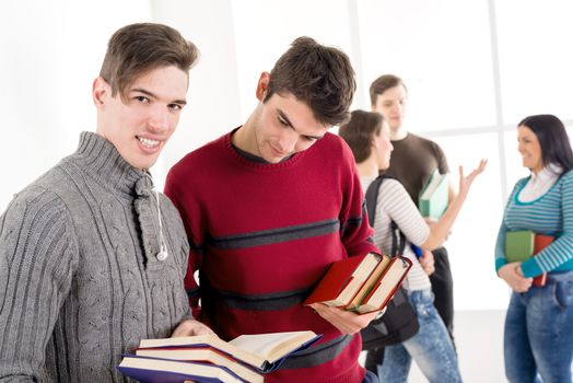 Two man student with book's is standing in the foreground and learning. A group of their friends is talking behind them.