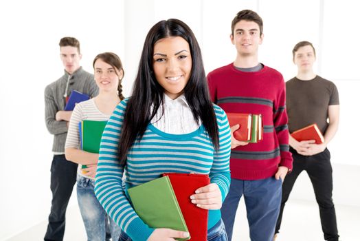 A beautiful smiling girl with book is standing in the foreground. A group of her friends is behind her. Looking at camera.