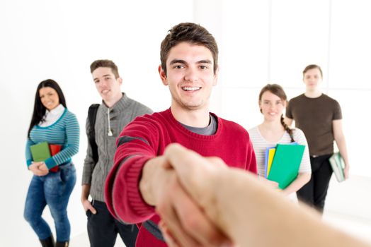 A young sucessful man student with books is standing in the foreground and shaking hand. A group of his friends is behind her. Looking at camera.