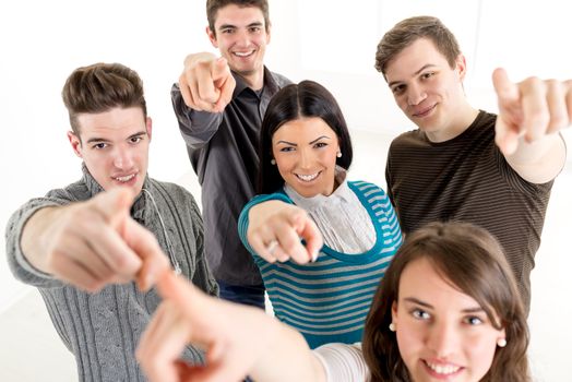 Group of cheerful students with book's standing and pointing with finger on camera in school hall and looking at camera.