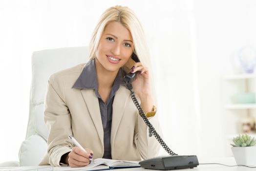 A young blond businesswoman phoning in office and with a smile on her face looking at the camera.
