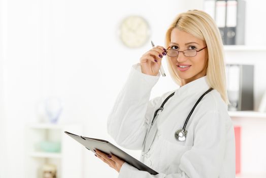 Young cute female doctor with the glasses, dressed in a white coat standing and holding folder with a stethoscope over the neck, with a smile looking at the camera.