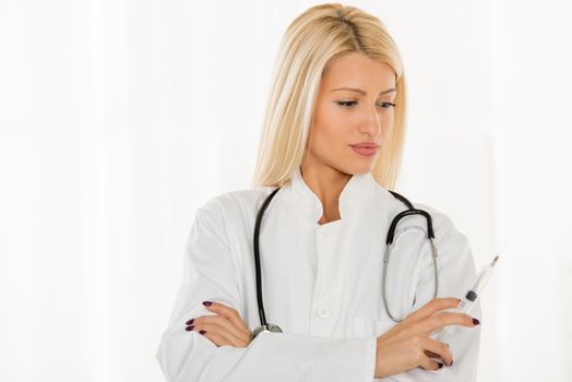 Pretty blonde young woman in a white coat, with a stethoscope over the neck holding an injection.