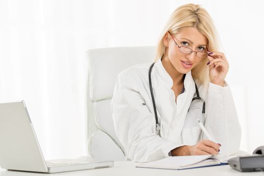 Attractive blonde female doctor in her consulting room, sitting at the table in front of laptop and looking at the camera, underneath her glasses.