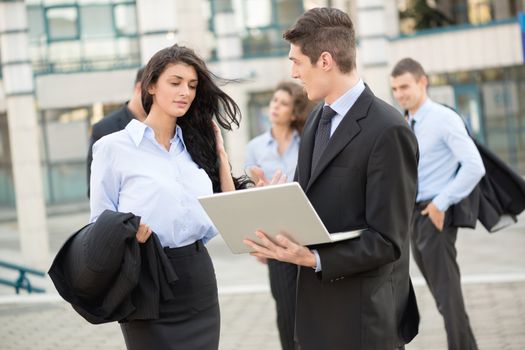 Young businessman talking to his business partner in front of office building holding a laptop while in the background see the rest of the business team.