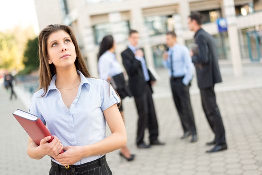 Young beautiful smiling businesswoman with planner in her hands, standing in front of office building separated from the rest of the business team. 