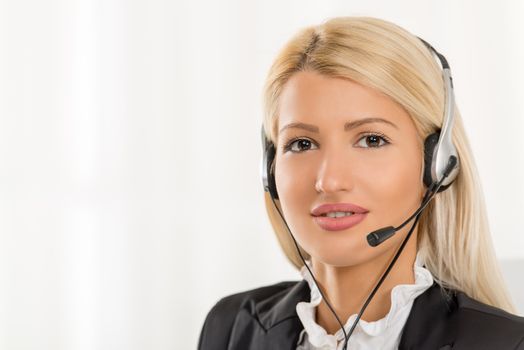 Close-up of a portrait young beautiful blonde girl, elegantly dressed, sitting at an office with a headset on her head. With a smile looking at the camera.