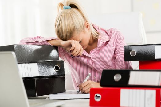 Young beautiful exhausted businesswoman sitting at a office desk between piles of binders and writes with her left hand.