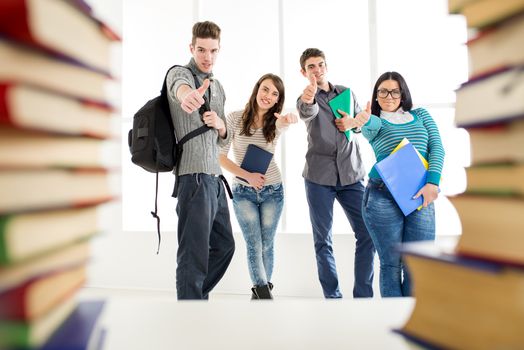 Group of cheerful students standing among the book's with thumb up in school hall and looking at camera.